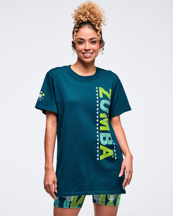 Teal short-sleeve crewneck T-shirt with vertical lime “ZUMBA” graphic, modeled by a female wearing patterned biker shorts on a white studio background.