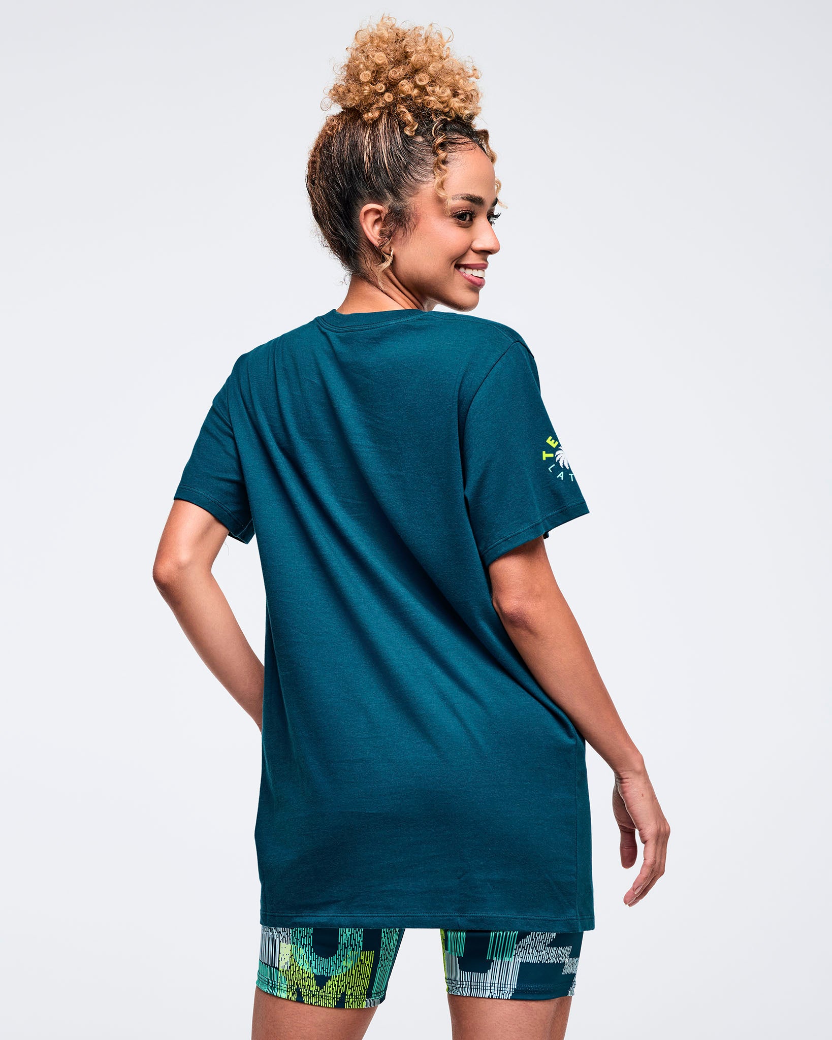 Back view of the teal crewneck T-shirt with short sleeves and a relaxed longline fit, modeled by a female wearing patterned biker shorts on a white studio background.