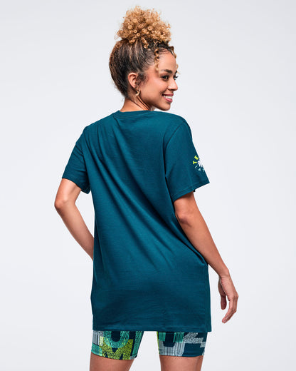 Back view of the teal crewneck T-shirt with short sleeves and a relaxed longline fit, modeled by a female wearing patterned biker shorts on a white studio background.