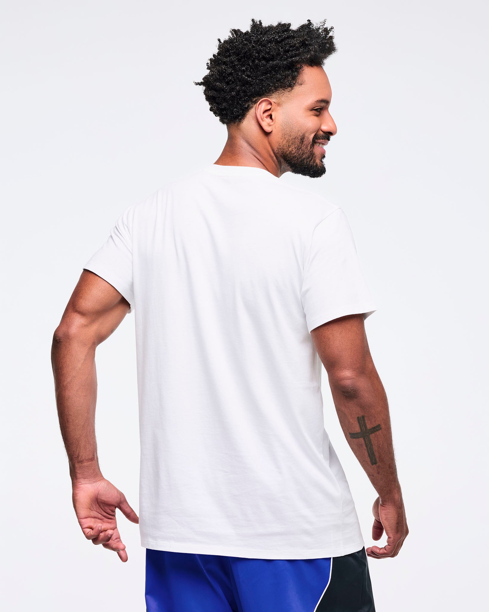 Back view of the white short-sleeve T-shirt on a male model, showing the clean solid back and relaxed fit, paired with bright blue track pants with black side panels and white piping.