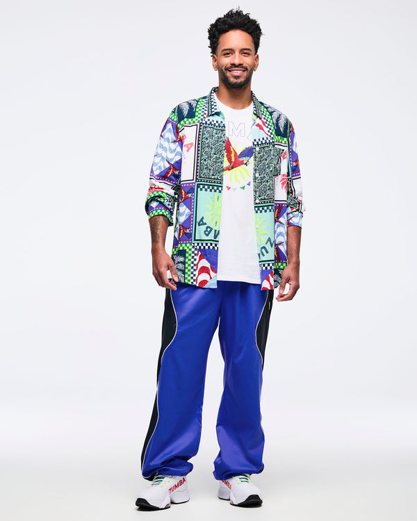 Full-body look featuring the white parrot graphic T-shirt layered under a long-sleeve button-up shirt with a multicolor patchwork print, worn by a male model with bright blue track pants and white sneakers.