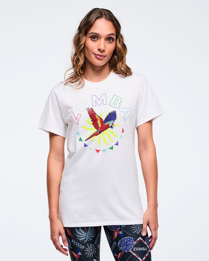White short-sleeve graphic T-shirt with a colorful parrot and sunburst print and “ZUMBA” lettering, worn by a female model with dark tropical-print leggings.