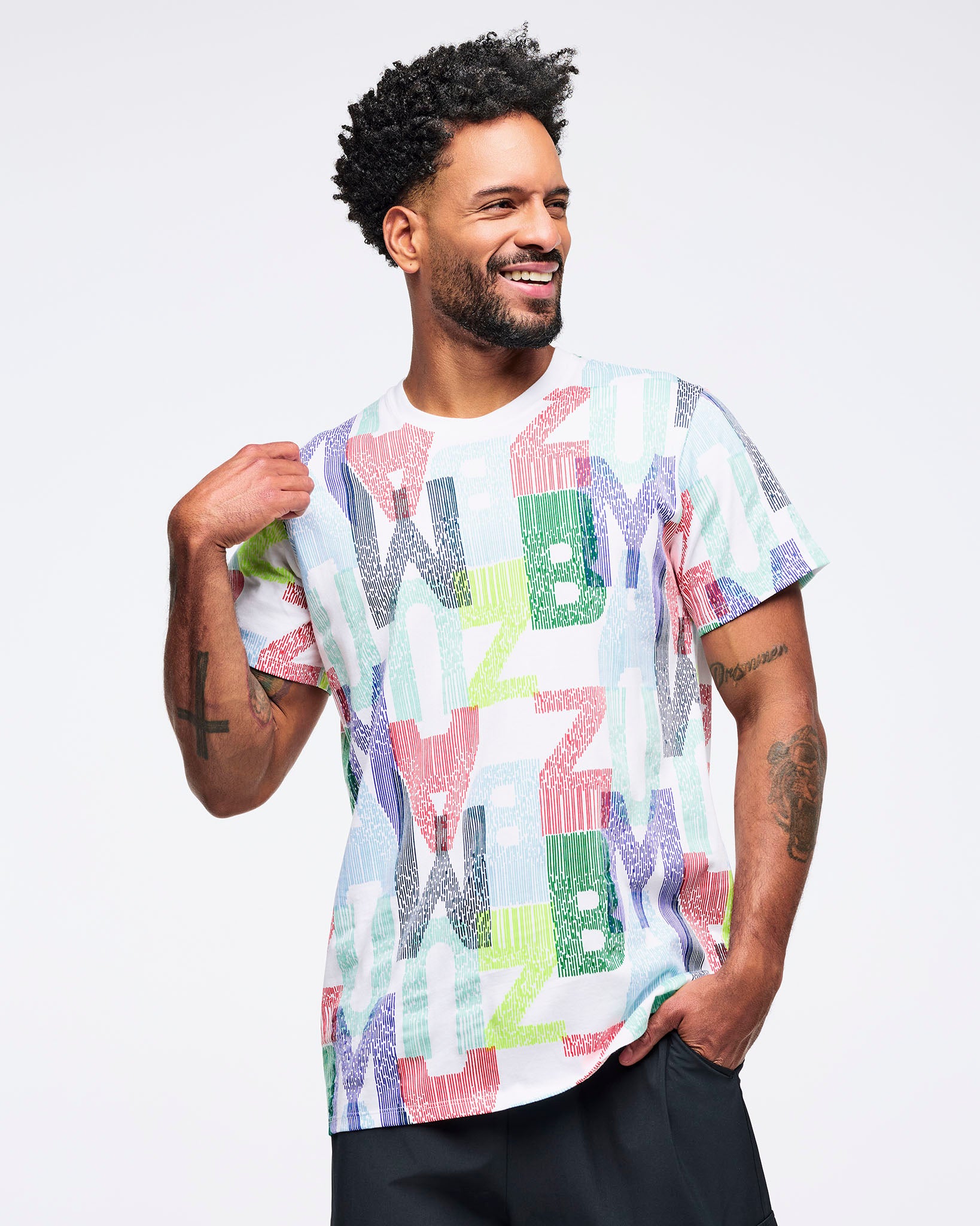 All-over print short-sleeve T-shirt with multicolor “ZUMBA” letter pattern, worn by a male model with black pants, posing with one hand in a pocket.