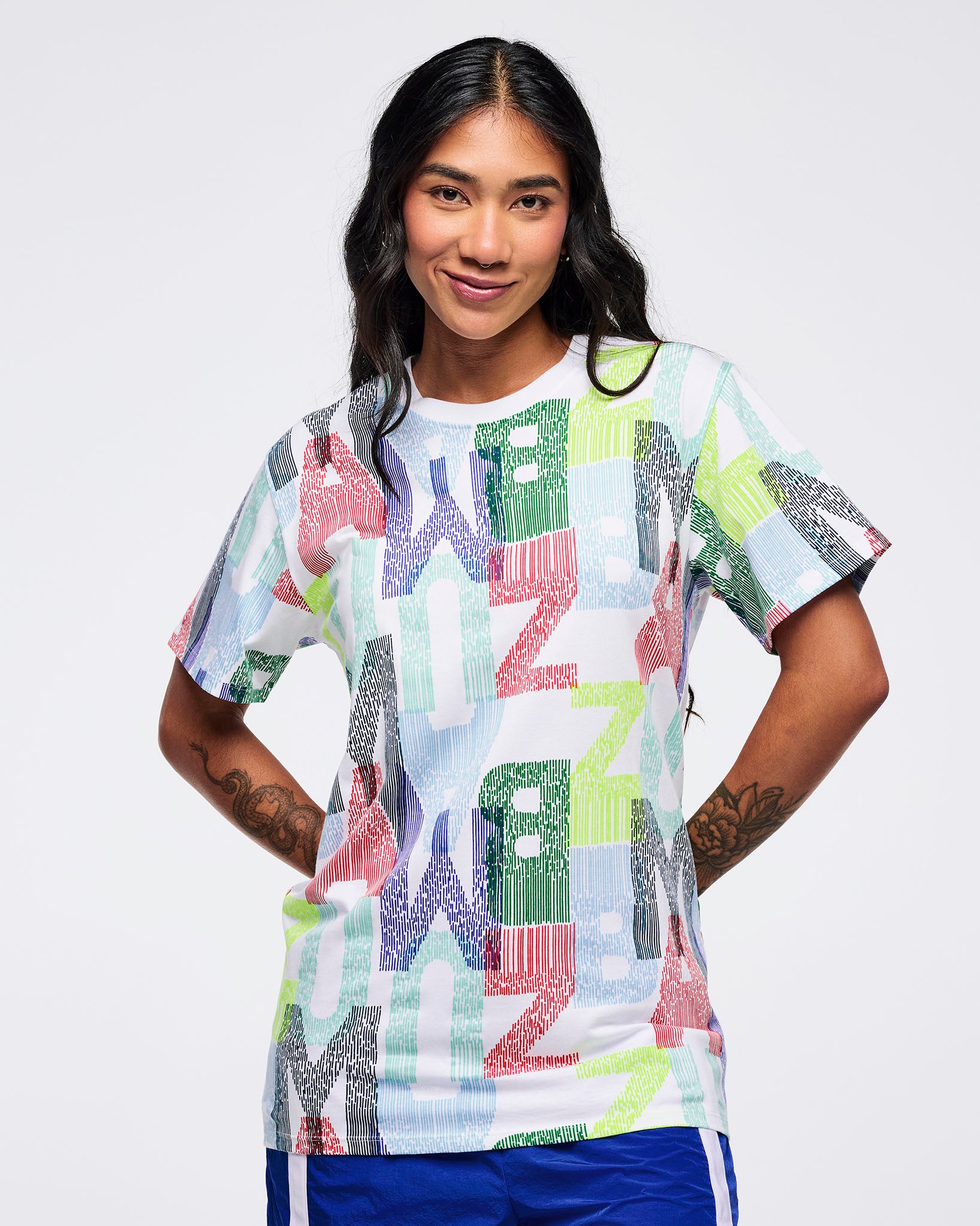 All-over print short-sleeve T-shirt with multicolor “ZUMBA” letter pattern on a white base, worn by a female model with bright blue track pants featuring white side stripes.