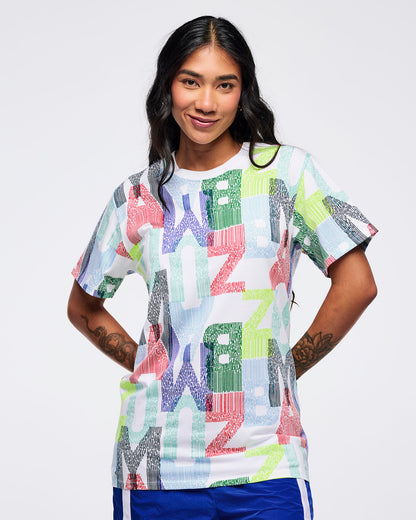All-over print short-sleeve T-shirt with multicolor “ZUMBA” letter pattern on a white base, worn by a female model with bright blue track pants featuring white side stripes.