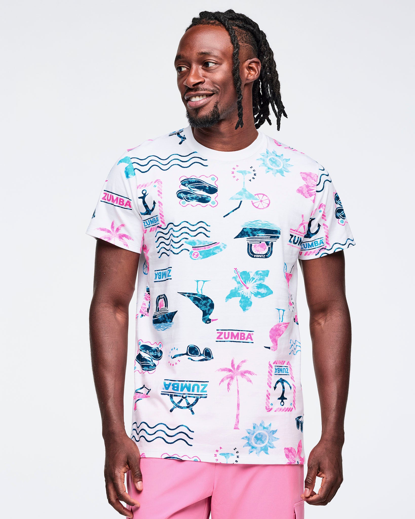 White short-sleeve crewneck Zumba graphic tee with an allover beach-themed print in teal and pink, including waves, anchors, palm trees, sunglasses, boats, and logos; shown on a male model with pink pants.