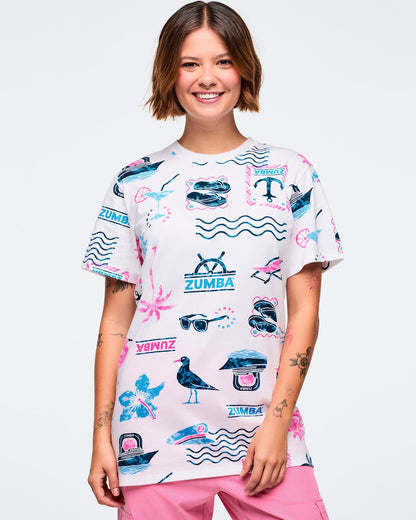 White short-sleeve crewneck Zumba graphic tee with an allover teal-and-pink beach print of waves, anchors, palm trees, sunglasses, boats, and logos; shown on a female model with pink pants.