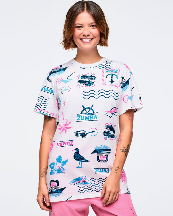 White short-sleeve crewneck Zumba graphic tee with an allover teal-and-pink beach print of waves, anchors, palm trees, sunglasses, boats, and logos; shown on a female model with pink pants.