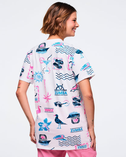 Back view of a white short-sleeve crewneck Zumba graphic tee with teal-and-pink allover nautical and tropical icons and logos; shown on a female model with pink pants.