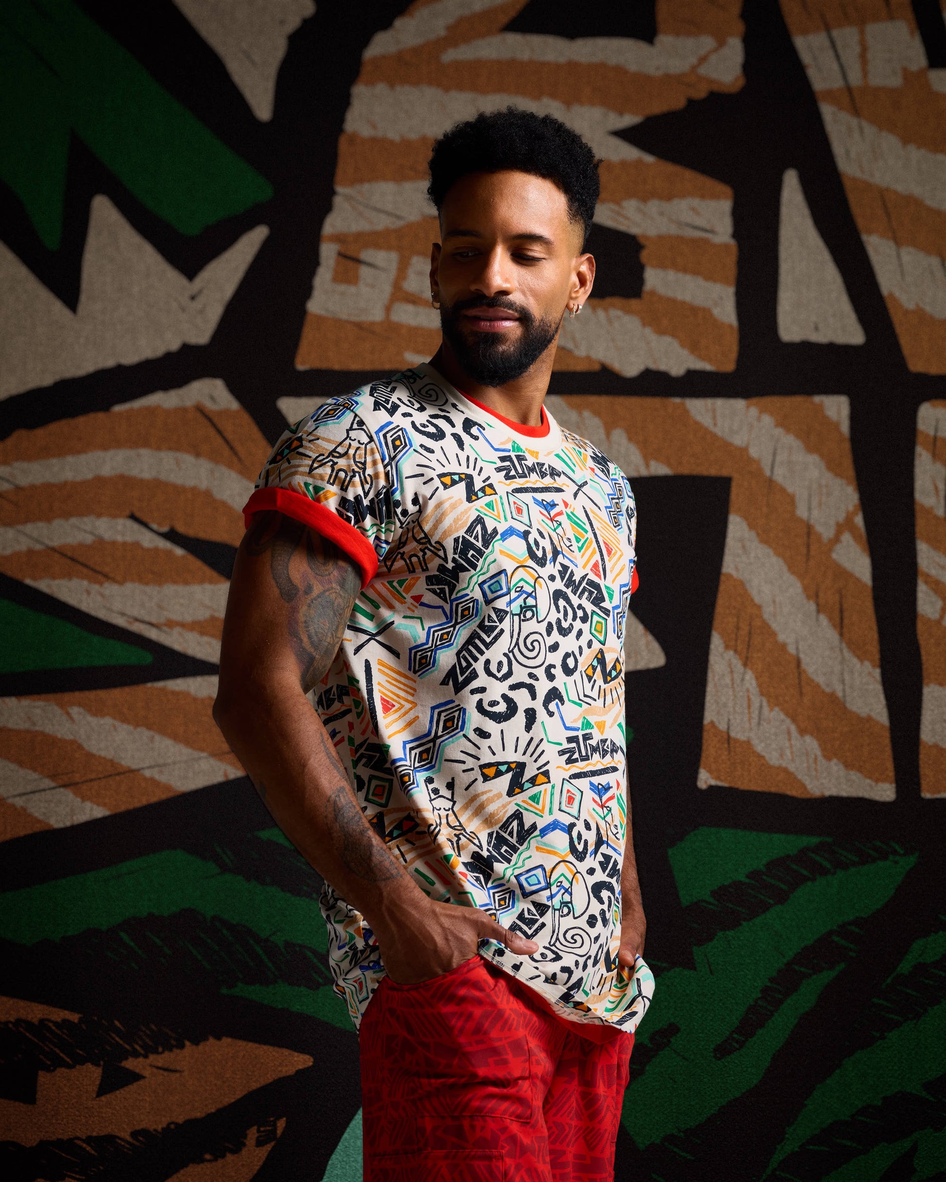 Male model in a tan graphic print tee with red trim and red cargo pants against a bold black, tan, and green backdrop.