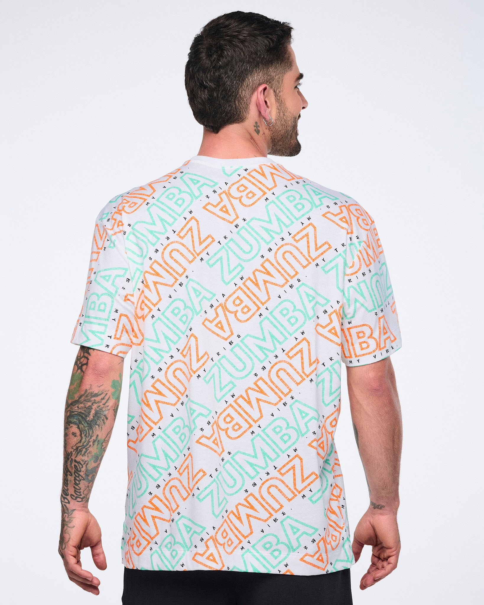 Back view of a white short-sleeve Zumba T-shirt with a crew neckline and allover mint and orange diagonal logo print. Male model wears the relaxed-fit tee with black pants.