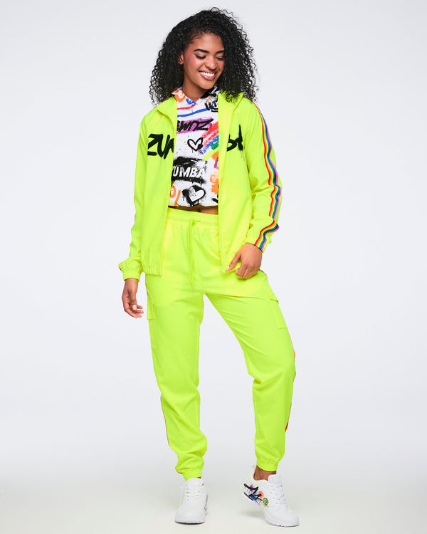 Female model wearing a bright yellow jacket with rainbow sleeve stripes over a colorful abstract hoodie, paired with bright yellow track pants featuring rainbow accents on a white background.