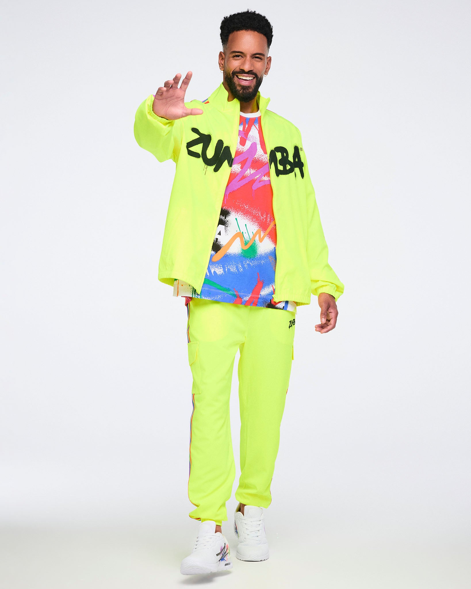 Male model wearing a bright yellow jacket over an abstract graphic T‑shirt, paired with bright yellow pants and white Zumba sneakers on a white background.
