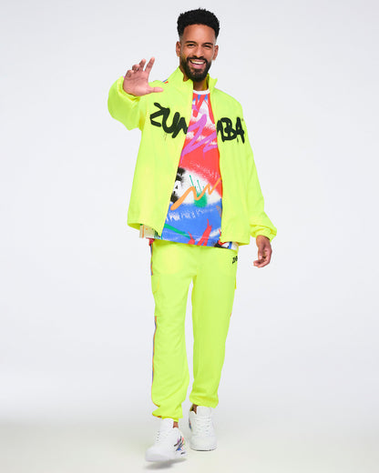 Male model wearing a bright yellow jacket over an abstract graphic T‑shirt, paired with bright yellow pants and white Zumba sneakers on a white background.