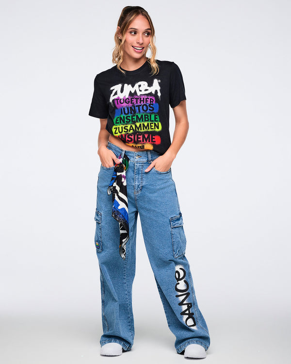 Female model wearing a black Zumba t-shirt with colorful text and denim cargo pants on a white background.