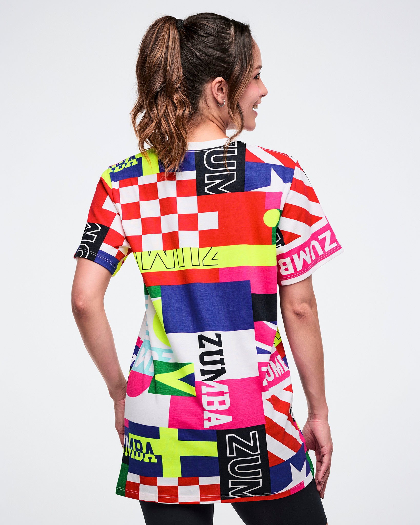 Back view of a female model wearing a colorful Zumba tee with various flag patterns against a white background.