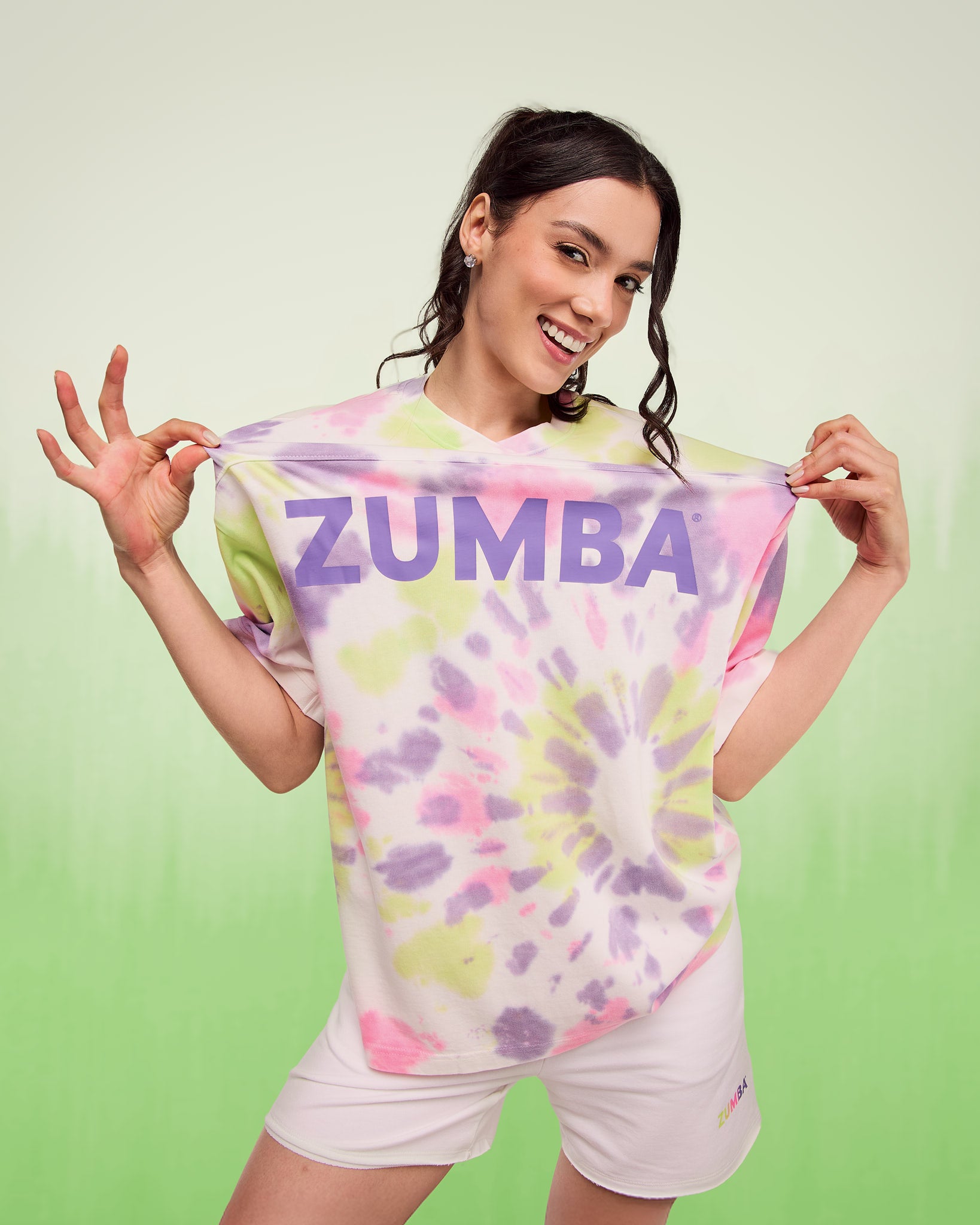 Smiling woman posing against a white-to-green gradient background, holding out the sides of a pastel tie-dye patterned “ZUMBA” T-shirt and wearing white shorts.