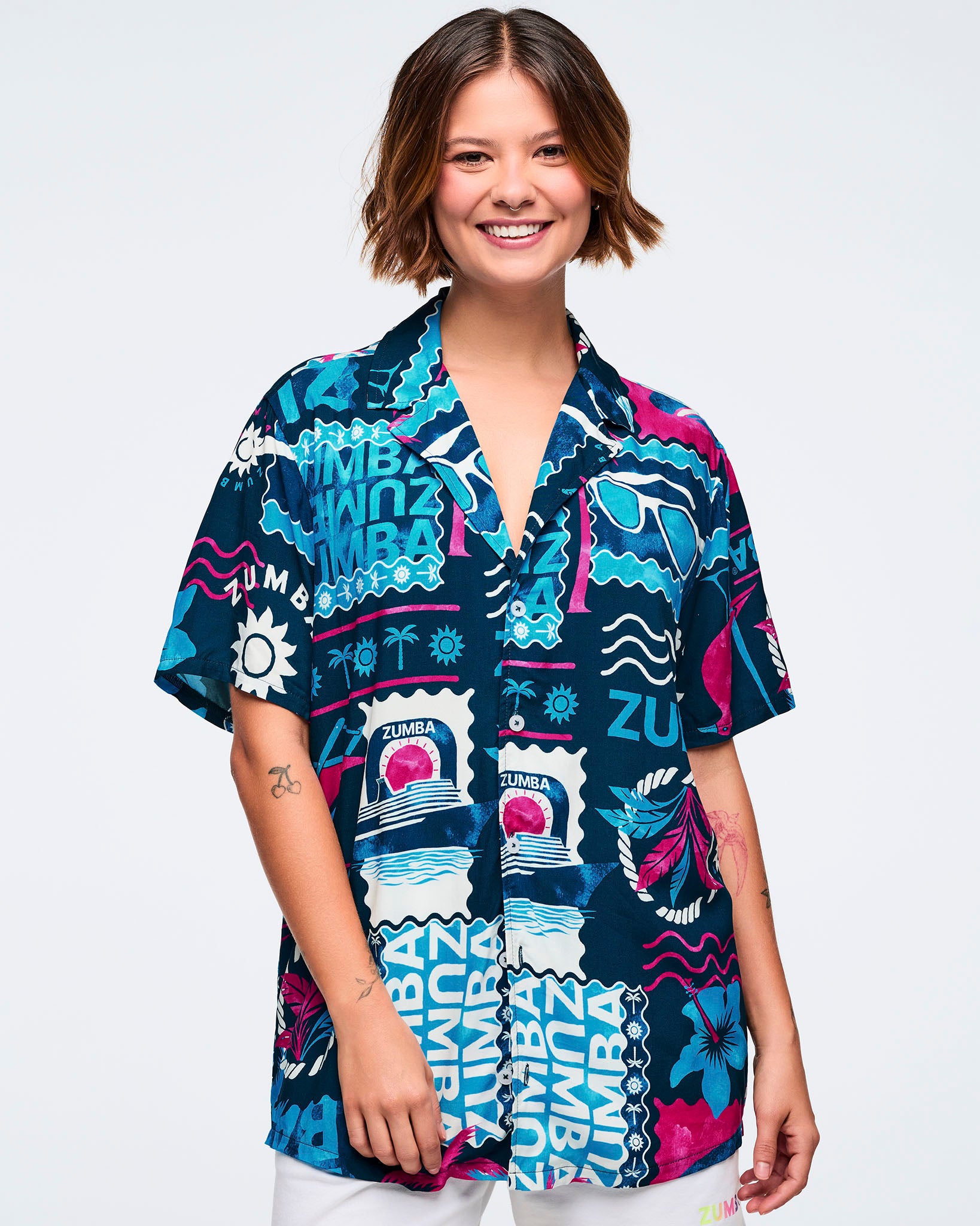 Navy short-sleeve button-up shirt with a relaxed collar and an all-over Zumba tropical print in teal, white, and hot pink featuring waves, palm trees, anchors, sunglasses, and logos; shown on a female model with white shorts.