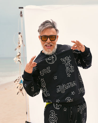 Beto, founder of Zumba, wears the coordinated black set sweatshirt and sweatpants, holding out the sweatshirt in front, near a beach backdrop.