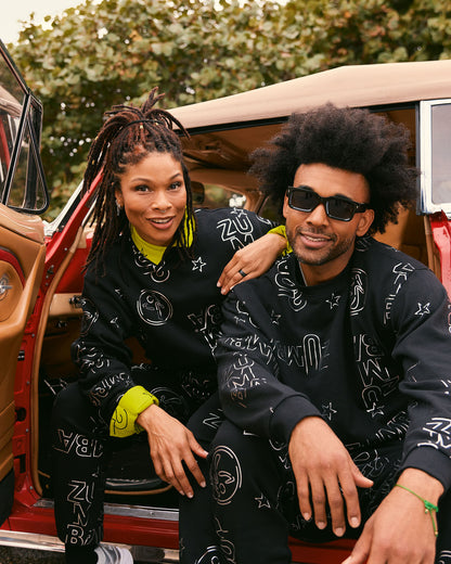 Black matching Zumba sweatshirt and jogger sets with white allover logo print. Female model and male model sit in the open door of a red vintage car wearing coordinated black sweatshirts and joggers layered with neon yellow button down worn underneath.