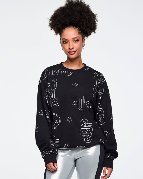 Black Zumba sweatshirt with an allover silver logo, star, and icon print. Female model is wearing the long-sleeve crewneck sweatshirt with shiny silver leggings featuring black side panels.