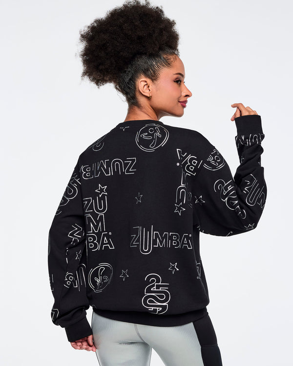 Black Zumba sweatshirt shown from the back with an allover silver logo, star, and icon print. Female model is turned away and wearing the long-sleeve crewneck sweatshirt with shiny silver leggings featuring black side panels.