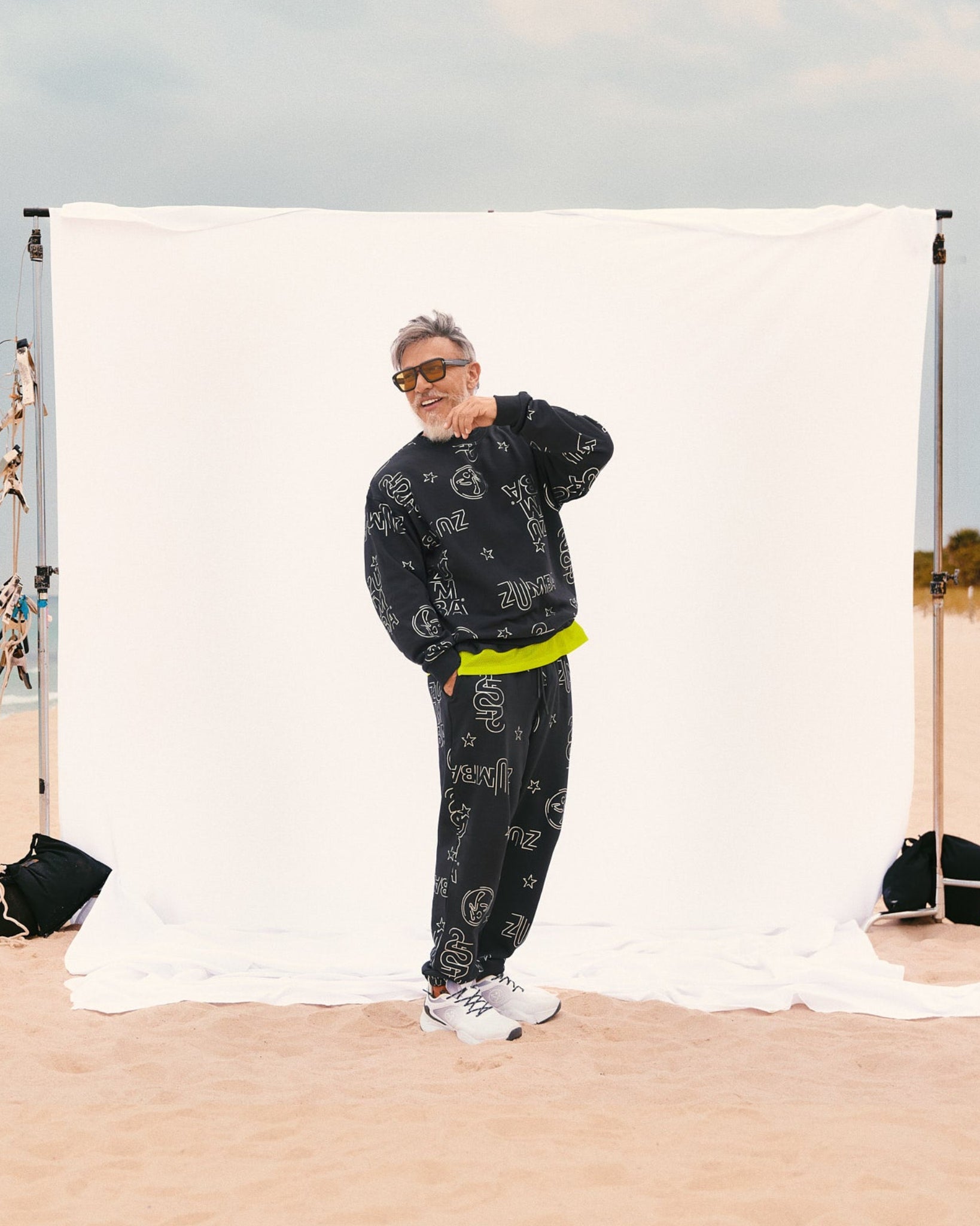 Beto, founder of Zumba, wears a relaxed-fit black crewneck sweatshirt and matching joggers layered over a Men's tee with an extended double layered mesh hem panel. Also wearing white sneakers, standing on sand in front of a white backdrop.