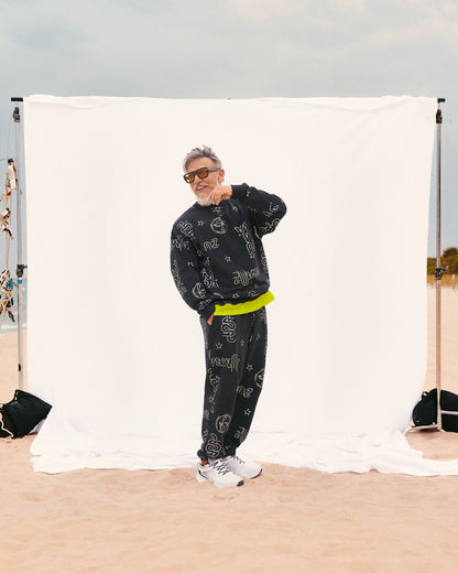 Beto, founder of Zumba, wears a relaxed-fit black crewneck sweatshirt and matching joggers layered over a Men's tee with an extended double layered mesh hem panel. Also wearing white sneakers, standing on sand in front of a white backdrop.