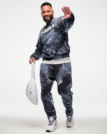Black and grey tie-dye crewneck sweatshirt with cream 'ZUMBA' text, paired with matching tie-dye joggers and a white 'ZUMBA'-print crossbody bag, male model.