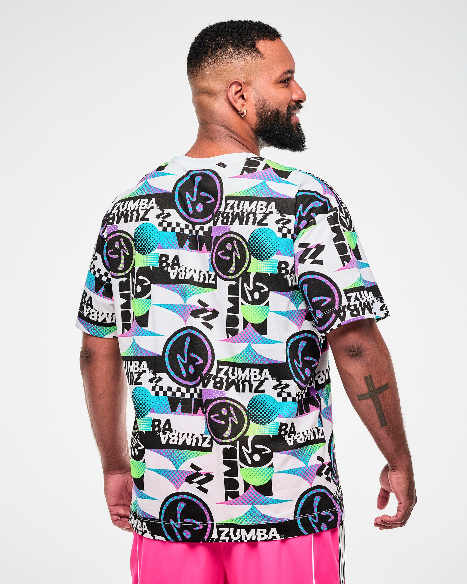 Back view of a male model wearing a white tee with a busy graphic pattern of repeated "ZUMBA" text and Zumbito logos.