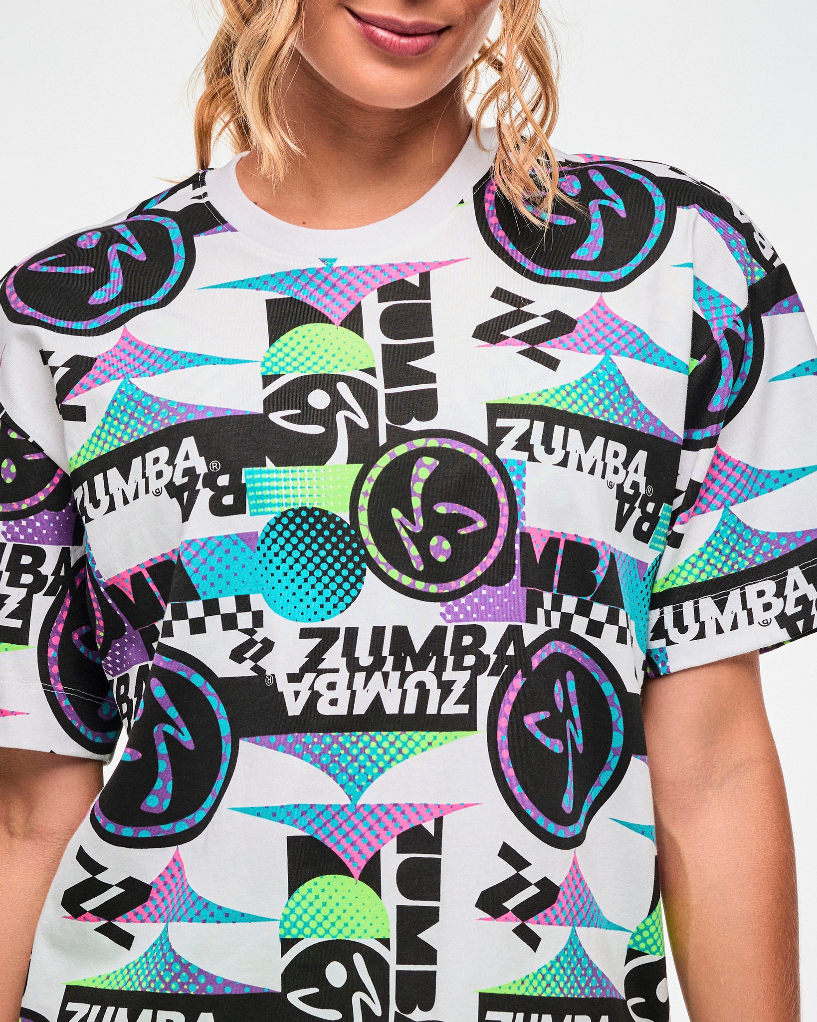 Close-up of a white tee with a busy black, purple, and aqua graphic pattern featuring repeated "ZUMBA" text and Zumbito logos.