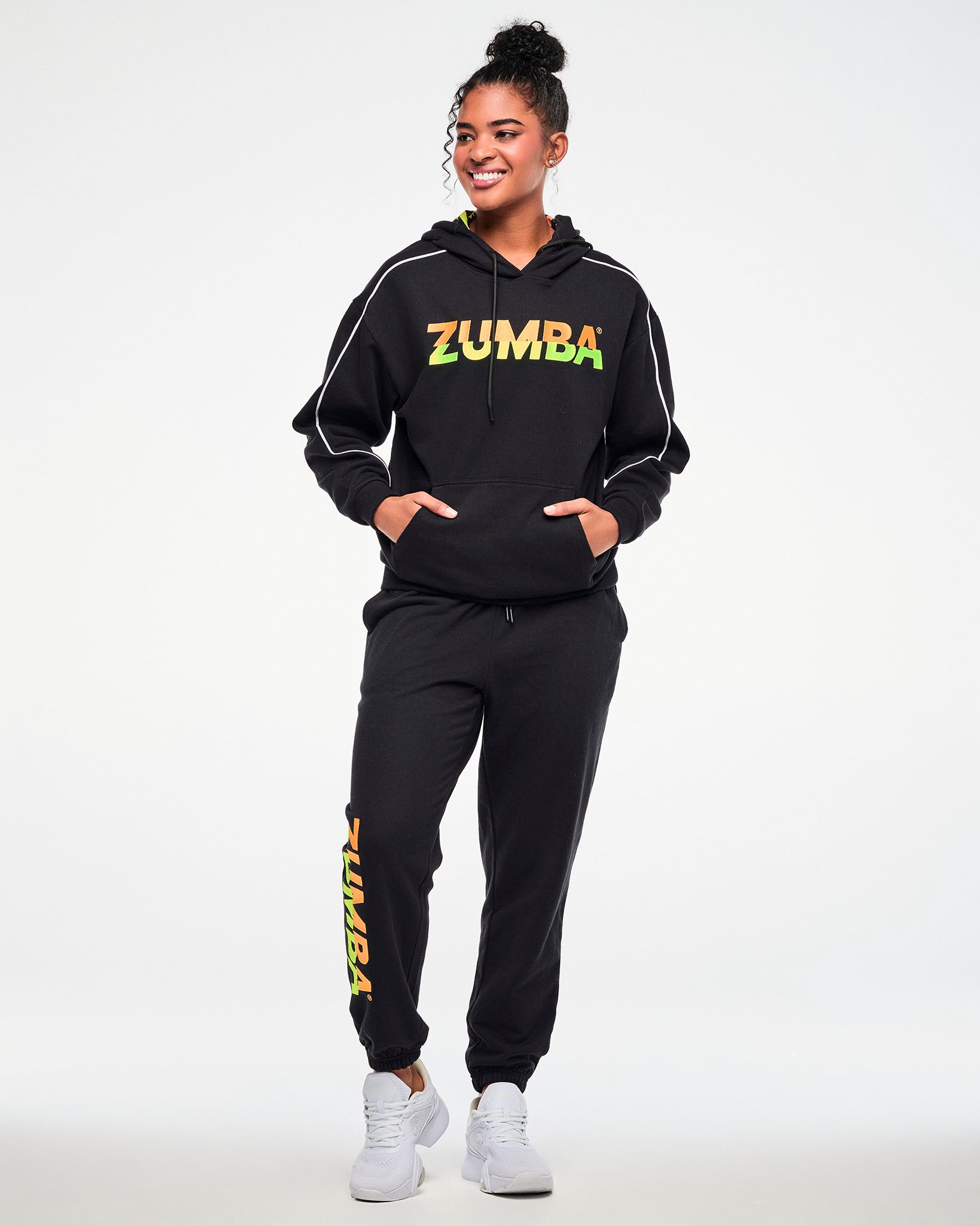 Female model wearing a black hoodie and matching joggers with a gradient "ZUMBA" logo, paired with white sneakers.