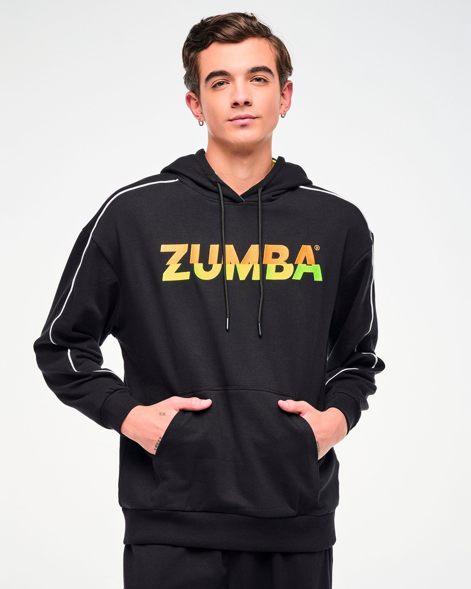 Close-up of a male model wearing a black hoodie with a gradient "ZUMBA" logo, shown with hands in the kangaroo hoodie pocket.