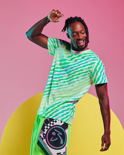Male model dancing and wearing a green and aqua lightning bolt striped tee with "ZUMBA" text, paired with neon green wide-leg pants with a graphic patch against a yellow and pink background.