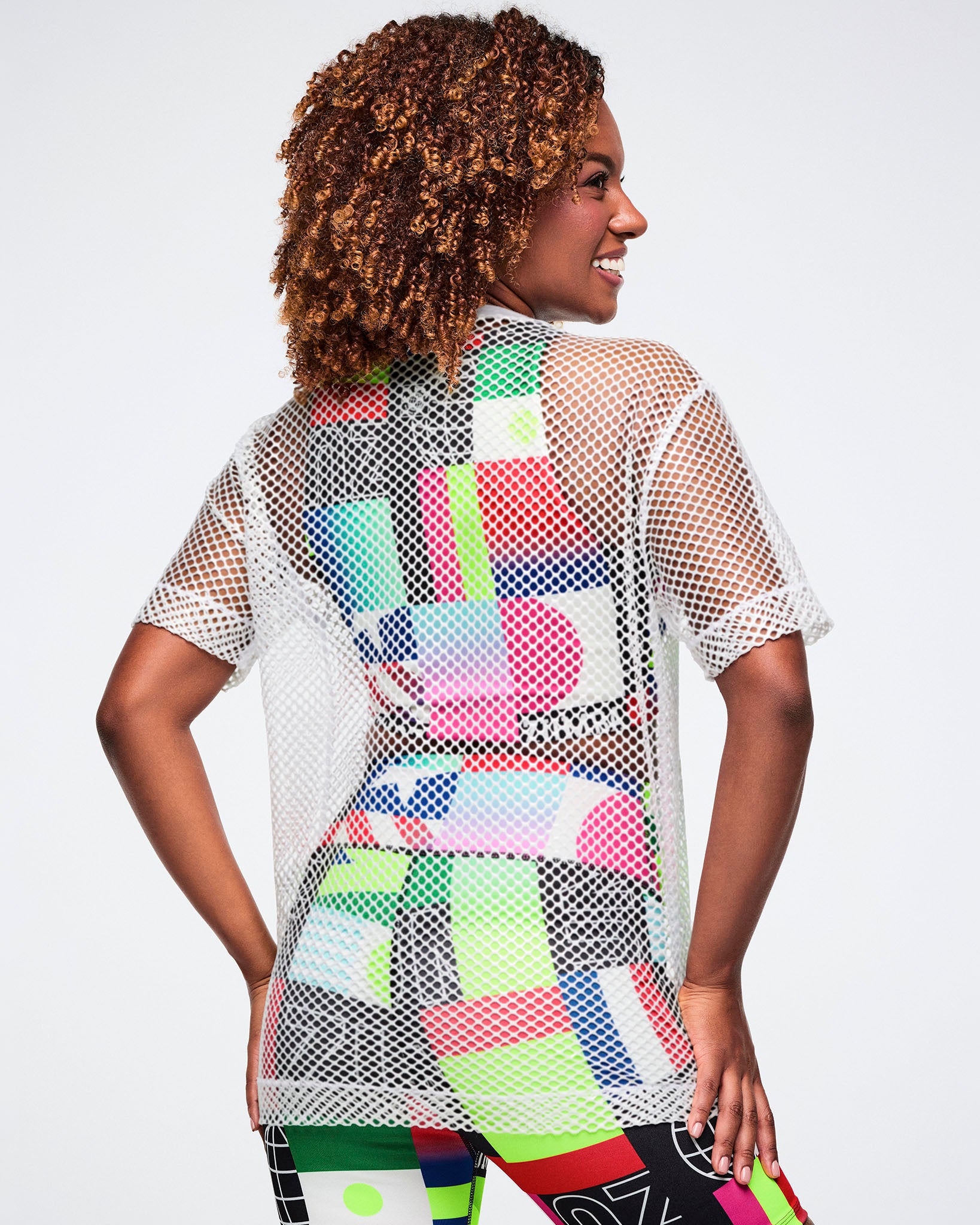 Back view of a female model in a colorful bra and biker shorts, layered with a white mesh top, set against a white background.