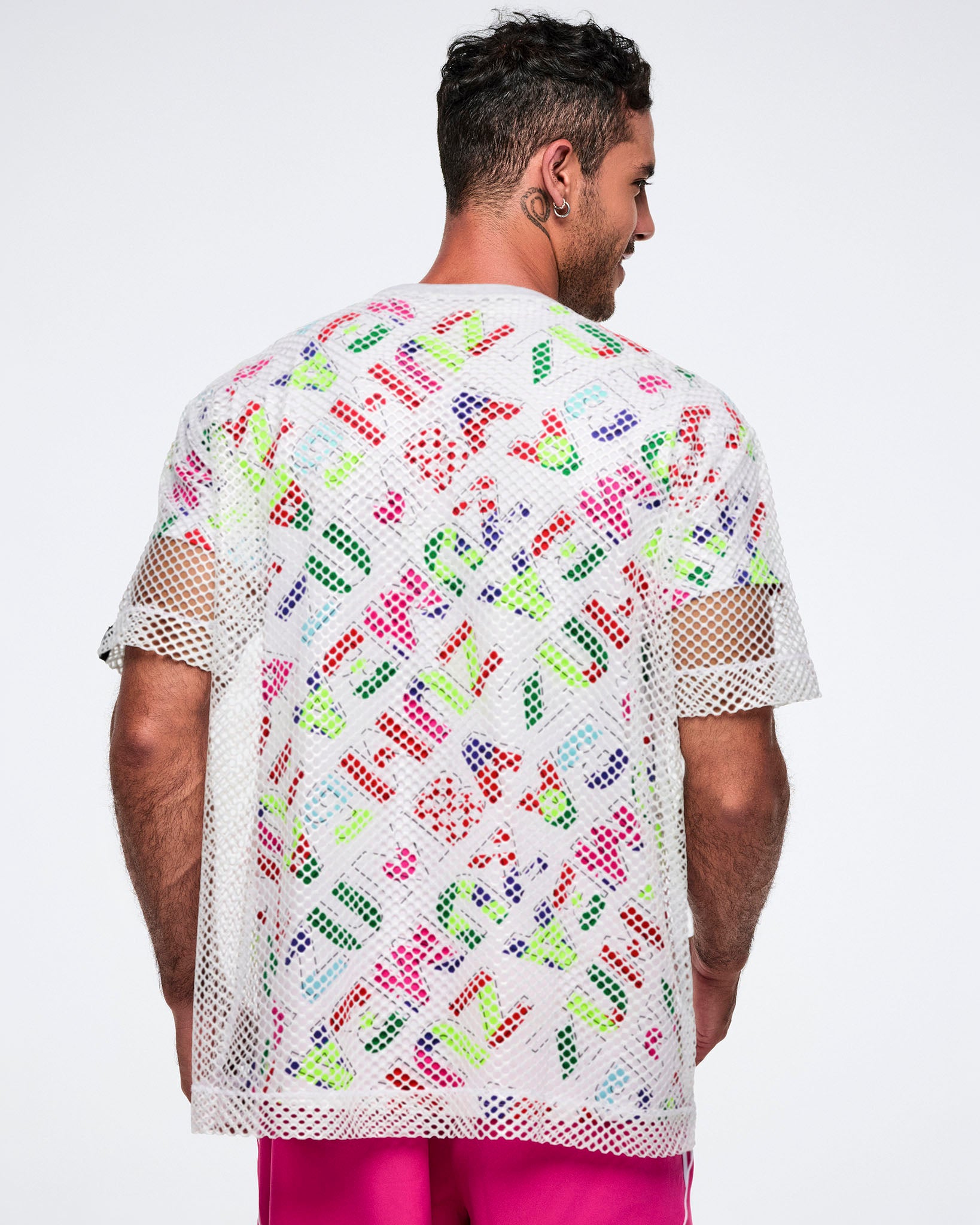 Back view of a male model wearing a colorful patterned shirt with a white mesh top layered over it, paired with pink pants.