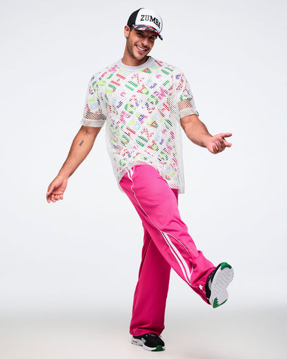 Male model posing with a forward kick, wearing a white mesh top over a colorful Zumba shirt, pink pants, black shoes, and a Zumba hat.