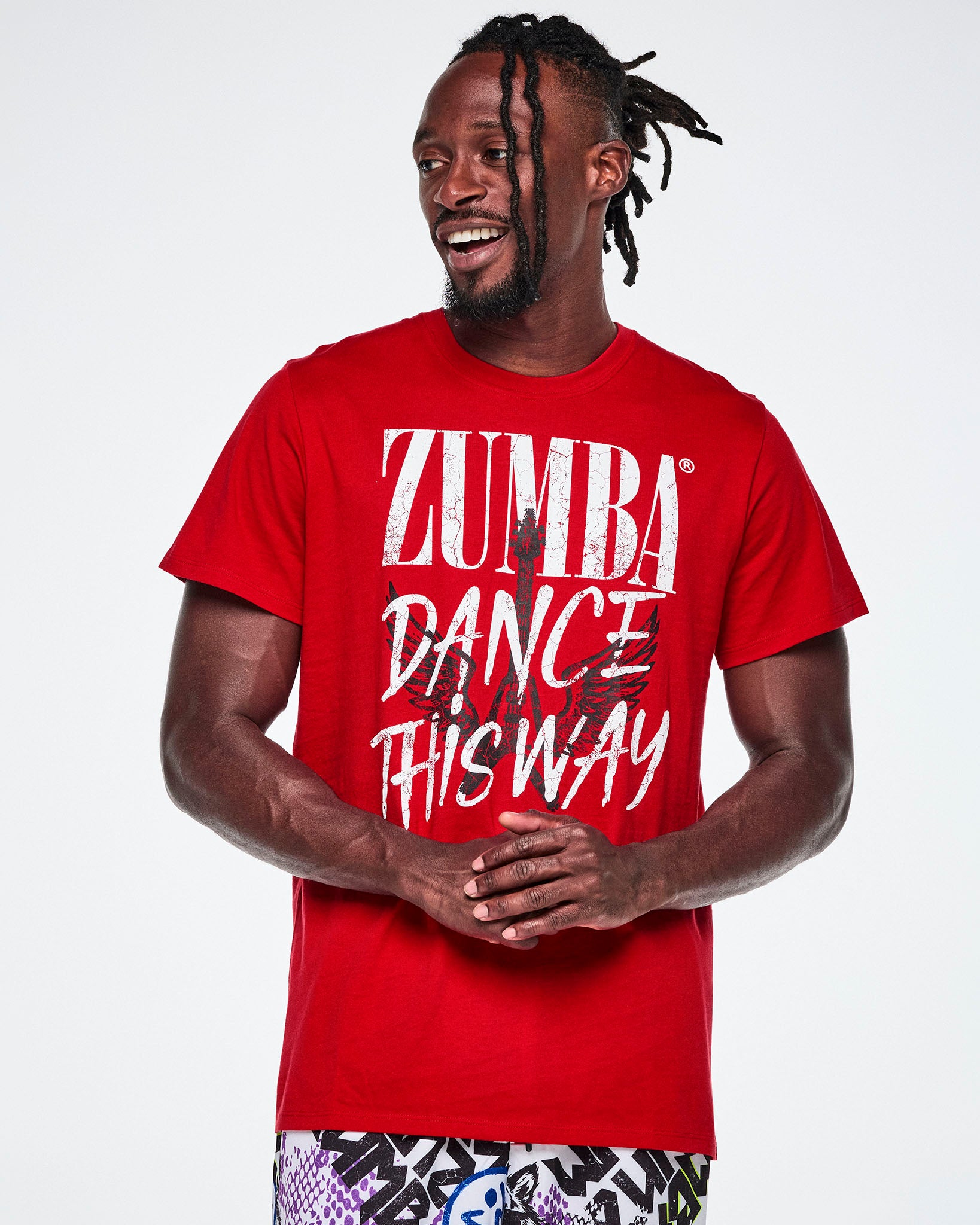 A male model wearing a red short-sleeve tee with a distressed white text that reads 'ZUMBA DANCE THIS WAY' with a black guitar and wings behind the text.
