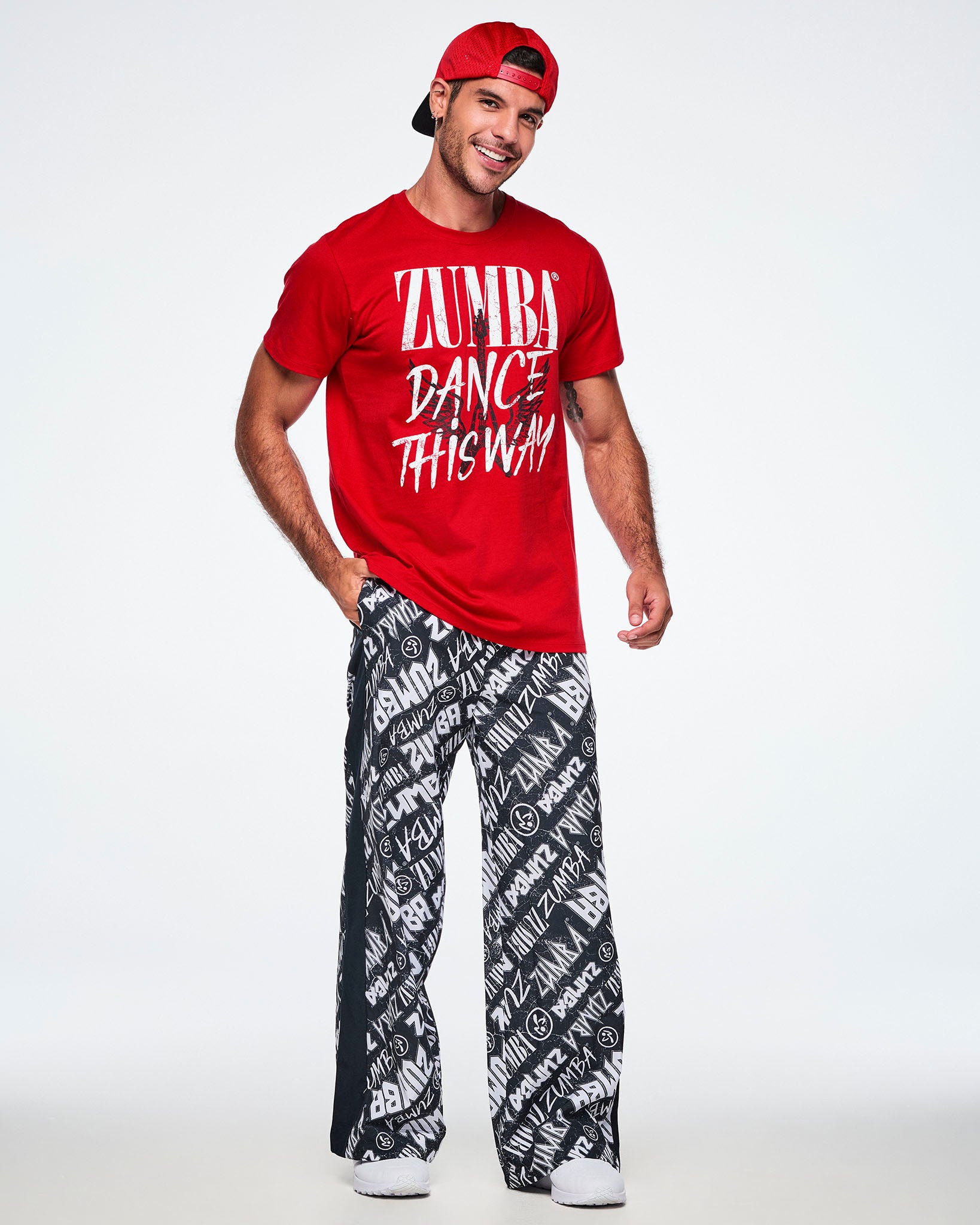 Full view of a male model wearing backwards red hat, a red short-sleeve tee with a distressed white text that reads 'ZUMBA DANCE THIS WAY' with a black guitar and wings behind the text and paired with black and white pants and white shoes.