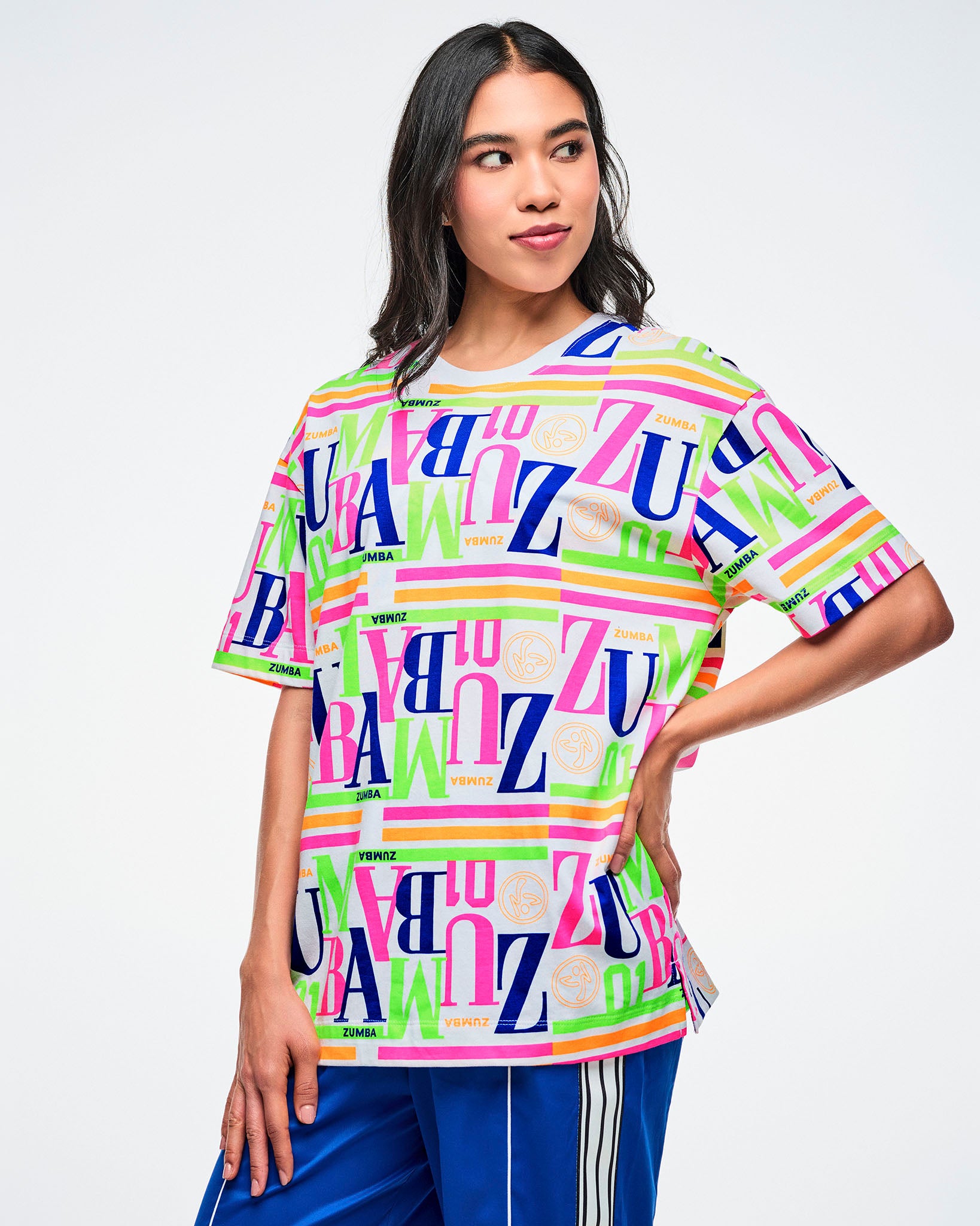 Female model wearing a colorful Zumba T-shirt with one hand at her side, paired with blue Zumba pants.
