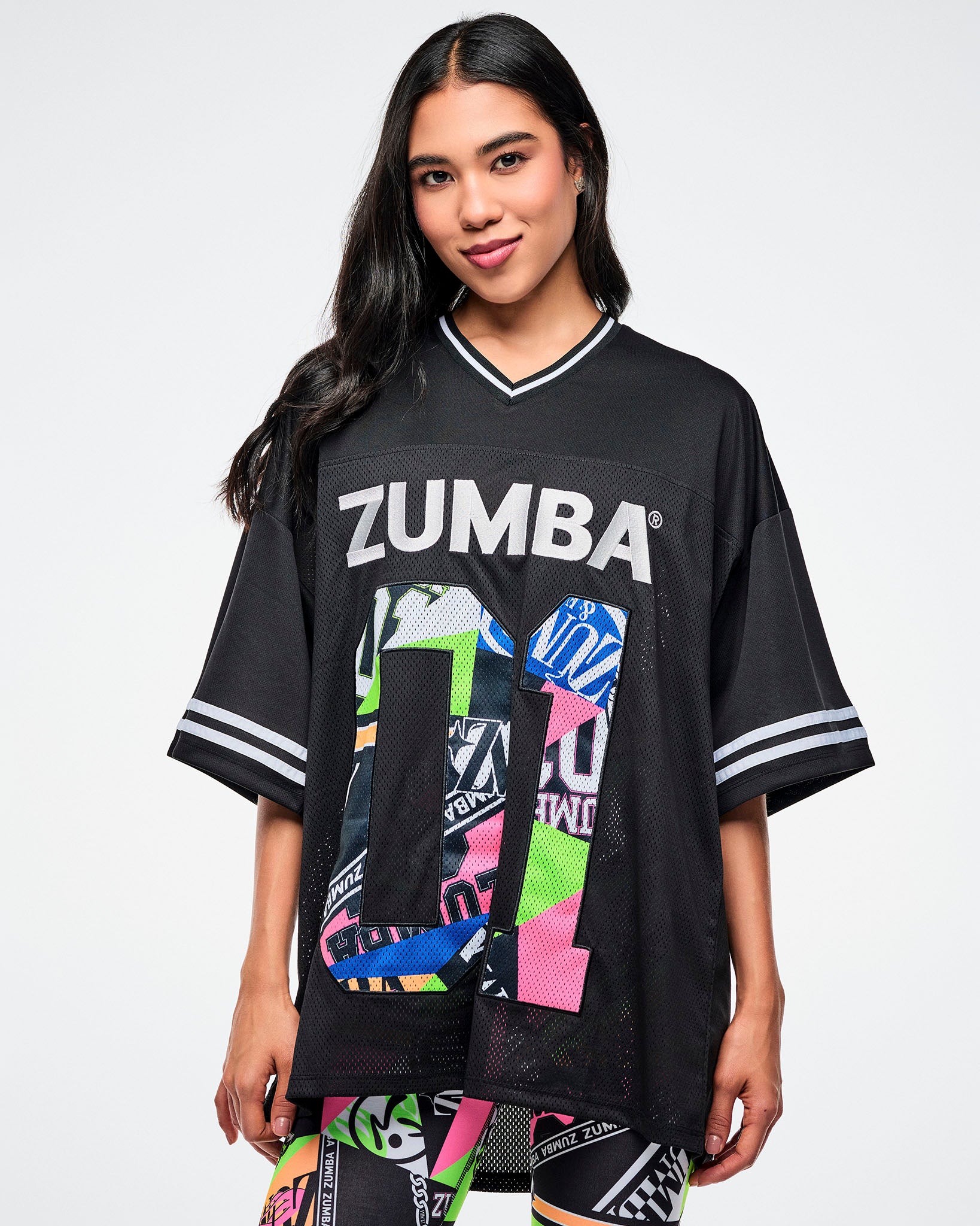 Female model wearing a black Zumba jersey with silver “ZUMBA” text and a large colorful patterned “01,” paired with matching patterned leggings on a white background.
