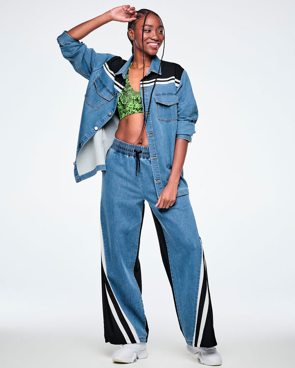 Female model wearing an open denim shacket with a black yoke featuring white stripes across the shoulders, layered over a neon green snake-print bra, paired with denim pants featuring a black and white striped side panel.