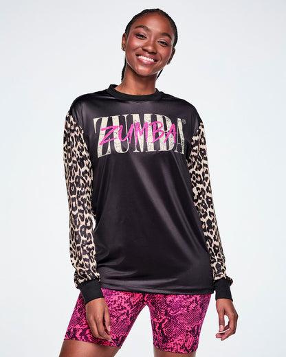 A female model wearing a black long-sleeve top with tan leopard-print sleeves and a pink and white Zumba text across the chest, paired with pink snake-skin patterned shorts against a white background.