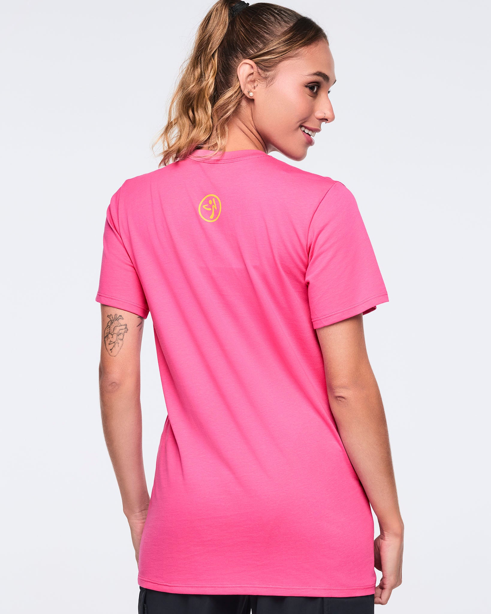 Back view of the bright pink short-sleeve Zumba T-shirt on a female model, featuring a small yellow, circular Zumbito logo printed below the back neckline.