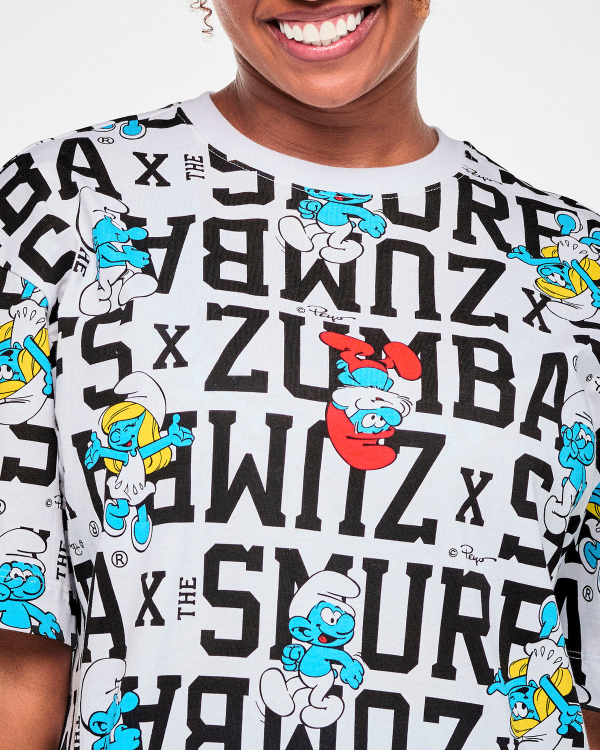 Close-up of the white top's all-over print: Papa Smurf in his red hat and pants, Smurfette with blonde hair and a white dress, and other blue Smurfs scattered among bold black 'ZUMBA x THE SMURFS' text.