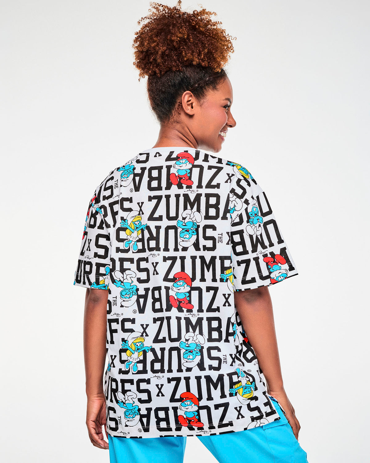 Shown from the back, a female model in a white oversized top with the all-over Smurfs print continuing across the back and sleeves, paired with aqua blue cargo pants.