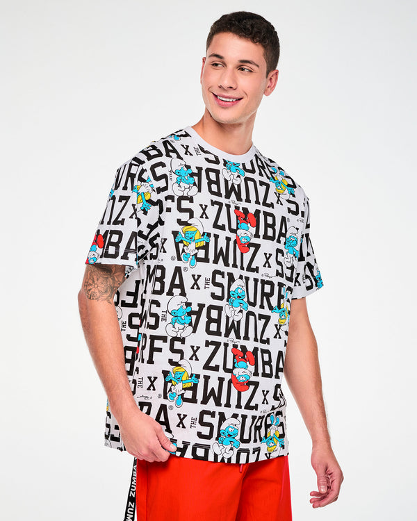Male model in a white oversized crew neck top with an all-over print of bold black varsity 'ZUMBA x THE SMURFS' text and Smurfs characters, including Papa Smurf and Smurfette, paired with red pants.