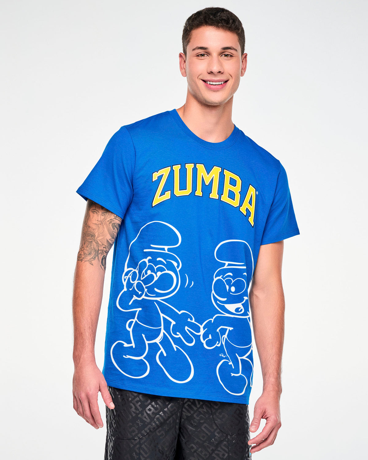 Male model in a blue crew neck tee with arched yellow 'ZUMBA' text above two white outline Smurfs dancing, paired with black pants with a tonal all-over logo print.