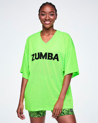 Female model wearing an oversized bright green mesh V-neck top with “ZUMBA” printed in black across the chest, paired with snake-skin patterned high-waisted biker shorts.