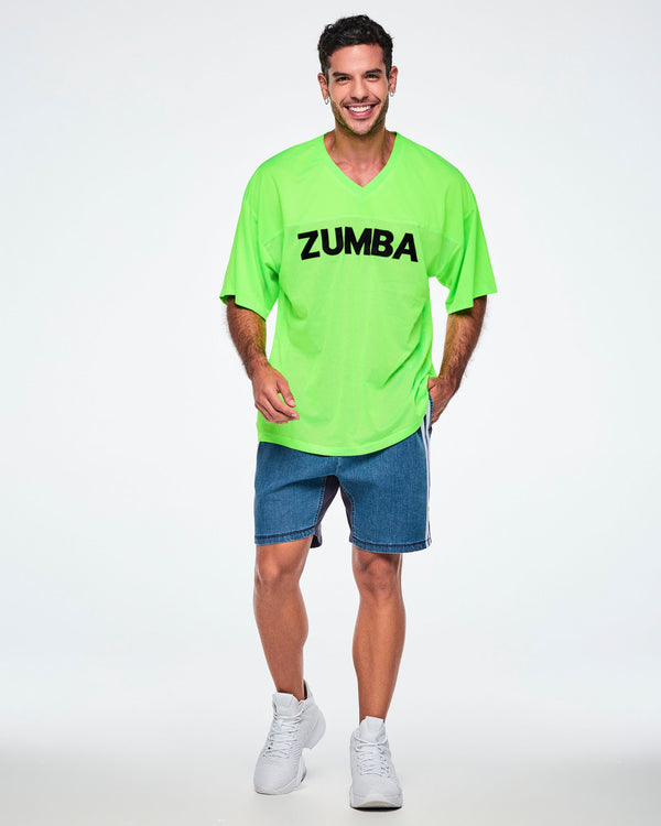 Male model with one hand in his pocket, wearing a neon green oversized V-neck jersey with a mesh body, solid shoulders and sleeves, and black 'ZUMBA' text across the chest. Paired with denim shorts with white side stripes.